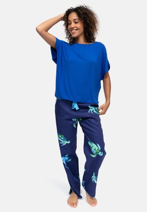 Donna sorridente con capelli ricci, indossa una maglia fluida blu e pantaloni blu navy con stampe di tartarughe marine, in piedi a piedi nudi su uno sfondo bianco.