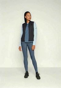 Fiatal nő sötétkék sport leggingsben, világoskék hosszú ujjú felsőben, fekete mellényben és fekete futócipőben áll, sima háttér előtt.