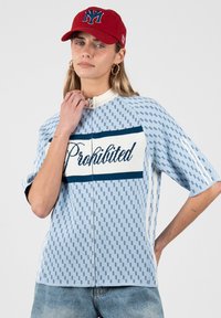 Femme portant une chemise zippée bleue à motifs avec le texte « Interdit » et une casquette rouge avec un logo bleu, posant avec une main près du cou.