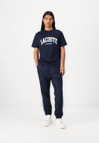 Jūras spēku Lacoste T-krekls ar baltu logo; kombinēts ar tumši ziliem sporta biksēm ar sānu svītrām. Baltas kedas noslēdz sportisko izskatu.