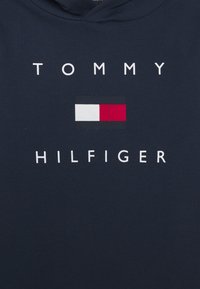 Granatowy sweter z widocznym białym napisem "TOMMY" oraz prostokątnym logo w czerwonych, białych i czarnych sekcjach poniżej "HILFIGER."