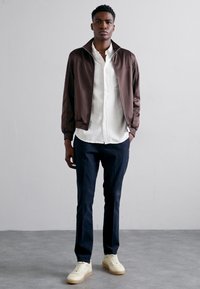Chaqueta bomber marrón sobre una camisa blanca, pantalones azul marino y zapatillas crema. Textura suave con puños y cuello acanalados. Diseño sencillo.