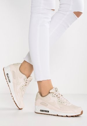 Osoba nosząca beżowe sneakersy Nike Air Max, zestawione z podartymi białymi obcisłymi dżinsami, stojąca na białej podłodze.