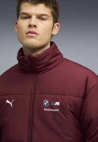 Veste matelassée bordeaux avec col haut, arborant le logo blanc de Puma et la marque BMW Motorsport. Texture lisse, fermeture zippée et détails cousus.