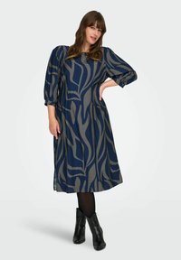 Vestido azul marino con una silueta fluida, que presenta rayas doradas diagonales y mangas abullonadas. Combinado con botas negras hasta el tobillo.
