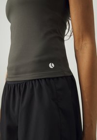Mørkegrønt tettsittende tanktop med et rundt logo, kombinert med løse svarte shorts. Jevn tekstur, minimalistisk design som fremhever komfort og bevegelighet.