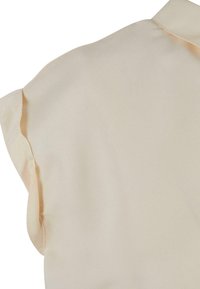 Beige ermeløs bluse med krage. Glatt, lett stoff, med subtile rynkede kanter langs ermehullene og halsutskjæringen.