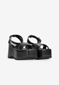Sandalias de plataforma negras con correas anchas cruzadas, correas ajustables en el tobillo y un tacón de cuña grueso. Fabricadas con un material similar al cuero suave.