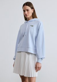 Femme portant un sweat à capuche bleu clair avec un logo et une jupe plissée blanche, debout devant un fond gris clair uni.