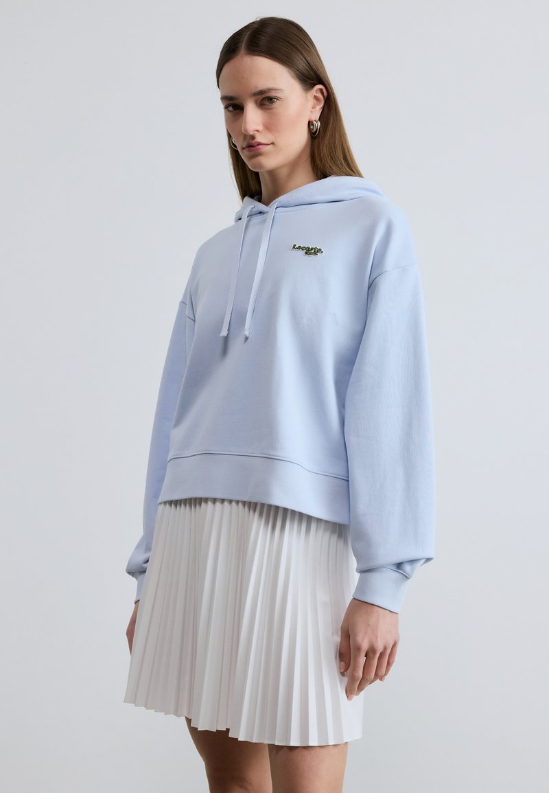 Femme portant un sweat à capuche bleu clair avec un logo et une jupe plissée blanche, debout devant un fond gris clair uni.