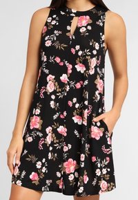 Schwarzes ärmelloses Kleid mit Blumenmustern in Rosa, Weiß und Braun. Es hat einen Schlüsselhalsschnitt und seitliche Taschen. Weicher, fließender Stoff.