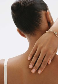 Pulsera de serpiente dorada en una muñeca, mano apoyada en un hombro desnudo. Textura de piel suave, cabello recogido en un moño ordenado. Tira de prenda blanca visible.