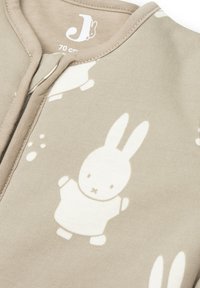 Combinaison en coton beige avec un motif de lapins blancs. Présente un col arrondi et une fermeture à pression sur le devant. Tissu doux et texturé. Étiquette de taille visible.