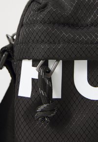 Bolso de tela negra con un patrón de diamante texturizado, que cuenta con un cierre superior, herrajes grises y una destacada impresión blanca del nombre de la marca.