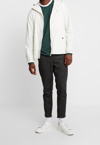 Biała lekka kurtka z kapturem, pod spodem zielony sweter o fakturze, ciemnoszare spodnie z zamkami przy kostkach oraz białe trampki.