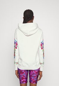 Persona con cabello trenzado que lleva una sudadera blanca con bordados florales coloridos en las mangas y pantalones cortos morados con estampado, mirando hacia atrás.
