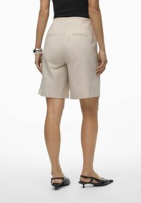 Pantalones cortos beige de corte ajustado con textura suave, pliegues delanteros y dos bolsillos traseros, combinados con zapatos de tacón negros.