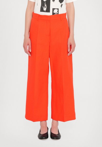 Bredde bukser i lysende orange stof med en glat tekstur og en høj talje. Stylet med sorte sko med lav hæl.