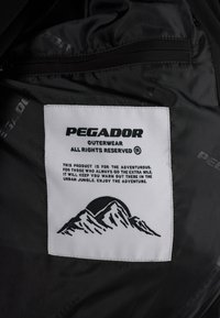 Svart stoffinteriør med en hvit etikett som har "PEGADOR OUTERWEAR" og et fjellgrafikk, som detaljert beskriver pleie og produktets formål.