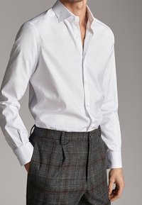 Man in een wit overhemd met knopen, ingestopt in grijs geruite broek, met ontspannen handen langs de zijkanten.