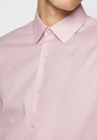 Camicia rosa chiaro con colletto a punte, dotata di bottoni perlacei e un tessuto morbido e liscio. Vista ravvicinata del collo.