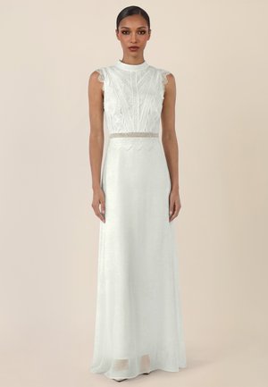 Frau steht, trägt ein ärmelloses weißes Spitzentop-Kleid mit hohem Ausschnitt und langem, fließendem Rock vor schlichtem Hintergrund.