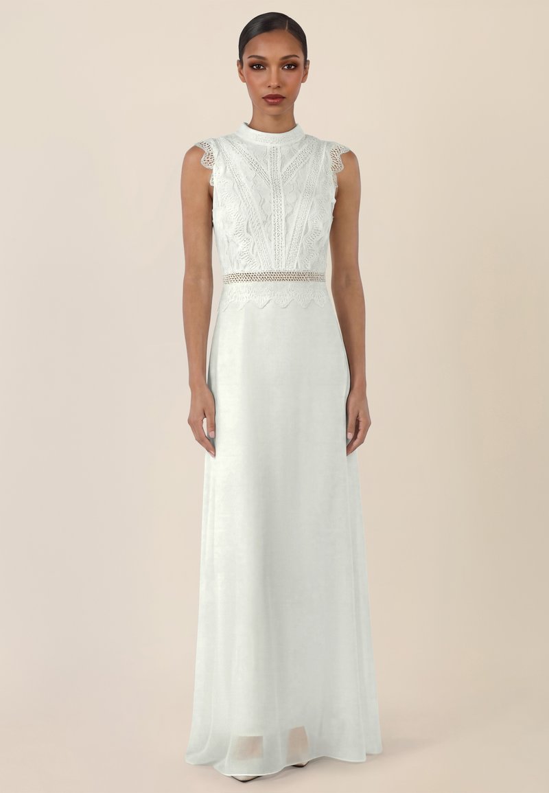 Vrouw staand, gekleed in een mouwloze witte kanten jurk met hoge halslijn en lange, vloeiende rok tegen een effen achtergrond.