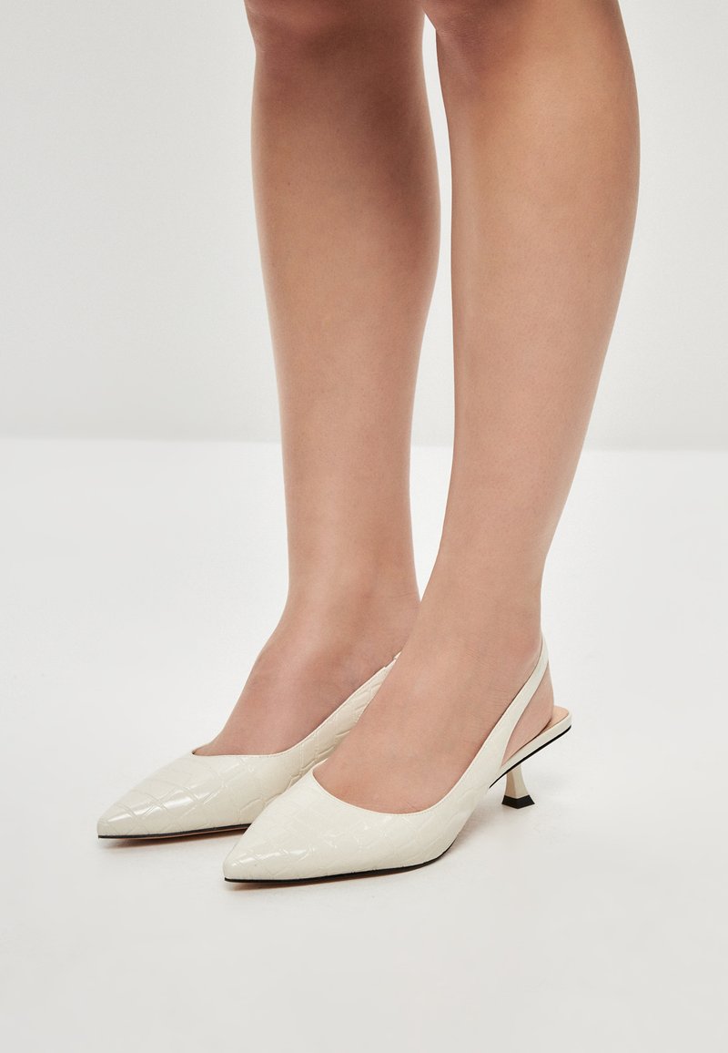 Valkoiset slingback-korkokengät, joissa on terävä kärki, kiiltävä pinta, pussitetttu rakenne ja lyhyt stiletto-korko. Varusteena on kontrastivärinen musta pohja.