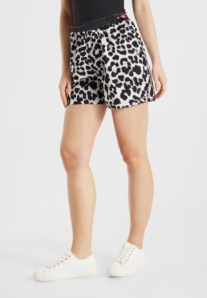 Kobieta w czarno-białych szortach w zwierzęcy wzór z czarną elastyczną talią i białych trampkach, stojąca na jednolitym tle.