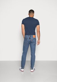 Camiseta de algodón navy combinada con jeans de denim azul que cuentan con un parche de cuero marrón y cinco bolsillos, usados con zapatillas blancas y navy.