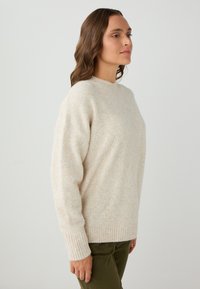 Pull en maille beige à encolure arrondie, avec des poignets côtelés et une texture douce. Associé à un pantalon de couleur olive, présentant une coupe décontractée.