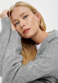 Sivý pletený sveter s rebrovanými manžetami, jemná textúra. Dlhé blond vlasy, prirodzený make-up, zlaté kruhové náušnice a uvoľnená póza.