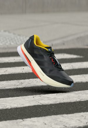 Černo-žlutá běžecká bota ve vzduchu nad mokrým přechodem pro chodce s malými kapkami vody stříkajícími z podrážky.