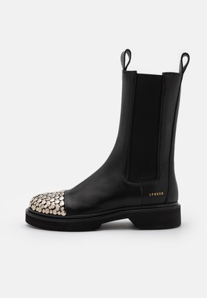 Bota de cuero negro hasta media pantorrilla con tiradores, paneles laterales elásticos, tachuelas circulares plateadas en la puntera y un pequeño texto dorado "CPNHGN" cerca del talón.
