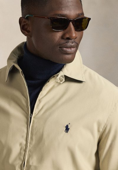 Casaco bege com fecho de correr e logo bordado em azul-marinho, usado sobre uma camisola de gola alta azul-marinho escura. Óculos de sol escuros com armações estampadas completam o visual.