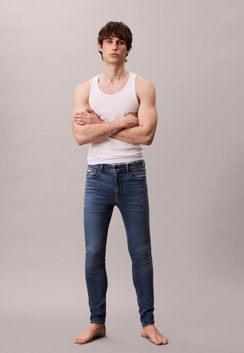 Fiatal férfi átfedett karokkal, mezítláb, fehér ujjatlan felsőben és karcsúsított kék farmernadrágban semleges háttér előtt áll.