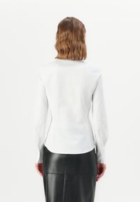 Blusa bianca aderente con maniche lunghe, texture liscia e orlo curvo, abbinata a una gonna a matita in pelle nera.
