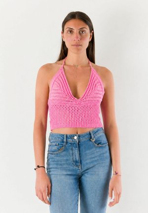 Donna che indossa un top corto rosa lavorato a maglia con spalline e jeans blu, in piedi davanti a uno sfondo bianco neutro.