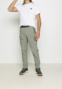 Lekki, oliwkowy model spodni trekkingowych o slim fit, z czarnymi akcentami, kieszeniami zapinanymi na zamek oraz czarnym paskiem, w połączeniu z białą koszulką i obuwiem outdoorowym.