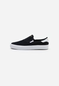 Svarta slip-on sneakers med vit sula och hältab, guldlogotyp på tungan och hälen, visade mot en enkel vit bakgrund.