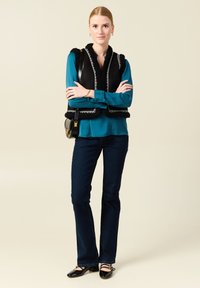 Gilet in finta pelliccia nera con cuciture bianche, blusa teal, jeans svasati blu scuro e scarpe nere con cinturini alla caviglia. Accessori minimi.