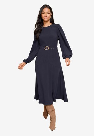 Tumši zilas krāsas kleita ar garām piedurknēm, pieguļošu augšdaļu un izplesto svārku. Tam ir dekoratīvs josts ar zelta sprādzi un gluda auduma tekstūra.