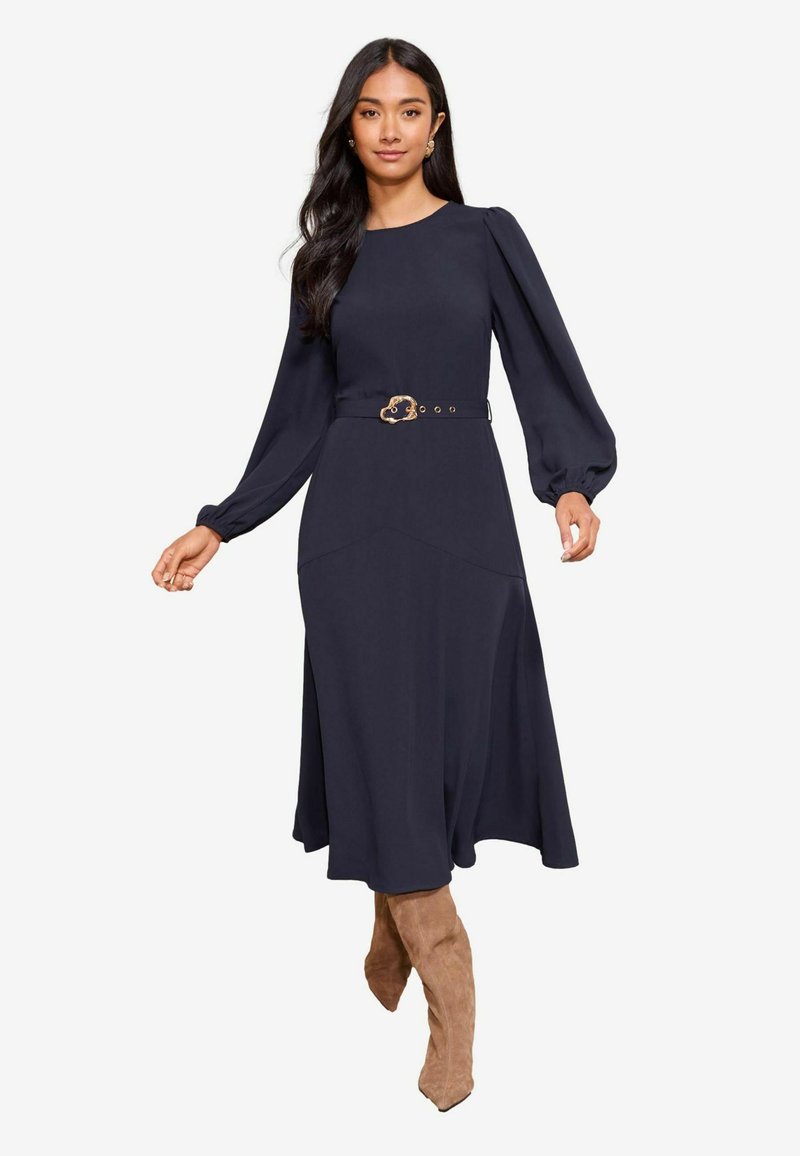 Tumši zilas krāsas kleita ar garām piedurknēm, pieguļošu augšdaļu un izplesto svārku. Tam ir dekoratīvs josts ar zelta sprādzi un gluda auduma tekstūra.