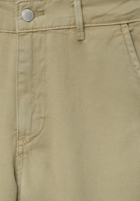Gros plan sur un pantalon en coton beige montrant la poche avant, la passants de ceinture, les coutures et une fermeture à bouton argenté.