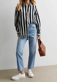 Mujer vistiendo una camisa de rayas verticales en blanco y negro, jeans azul claro, zapatillas blancas, sosteniendo un bolso marrón de lado frente a un fondo plano.