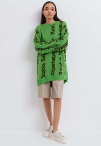 Groene oversized trui met zwarte stadsnamen bedrukt, gecombineerd met beige shorts en witte platform sneakers, tegen een effen achtergrond.