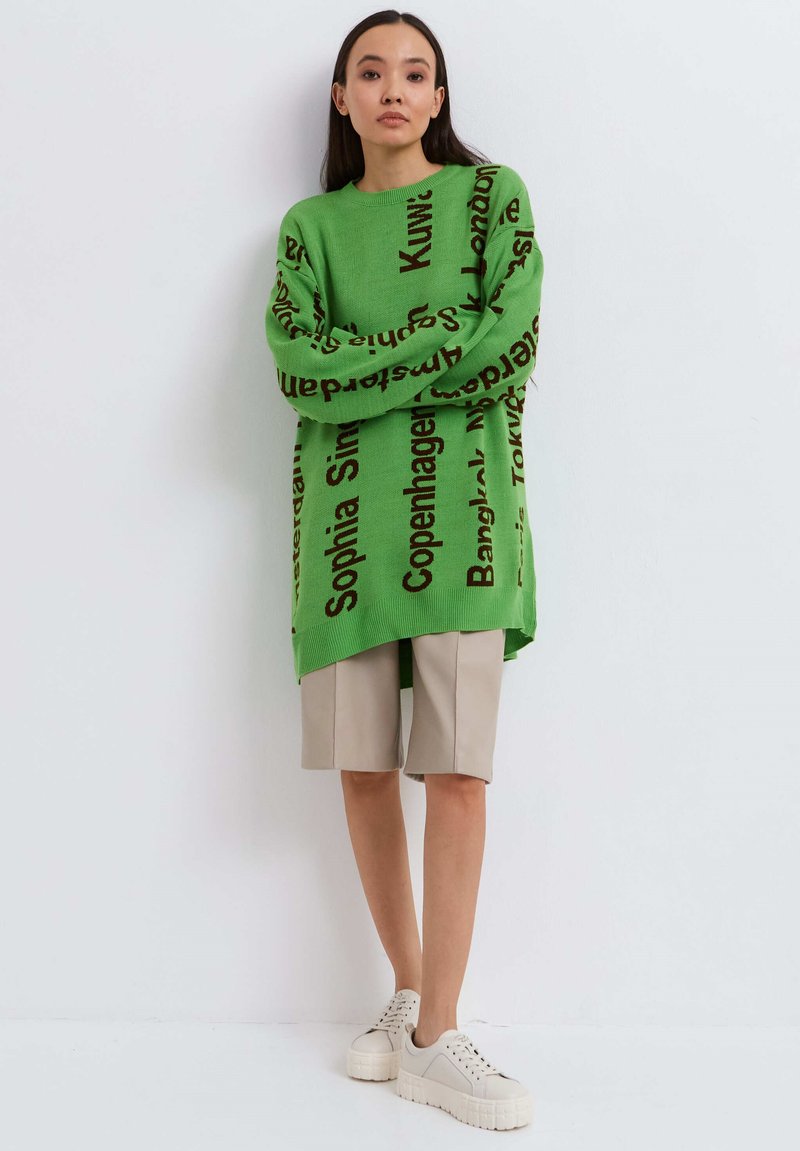 Groene oversized trui met zwarte stadsnamen bedrukt, gecombineerd met beige shorts en witte platform sneakers, tegen een effen achtergrond.