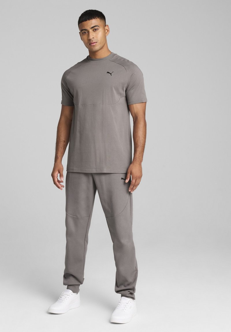 Šedý sportovní outfit skládající se z trička s krátkým rukávem a zúžených kalhot, s hladkým povrchem a vyšitým logem na hrudi.