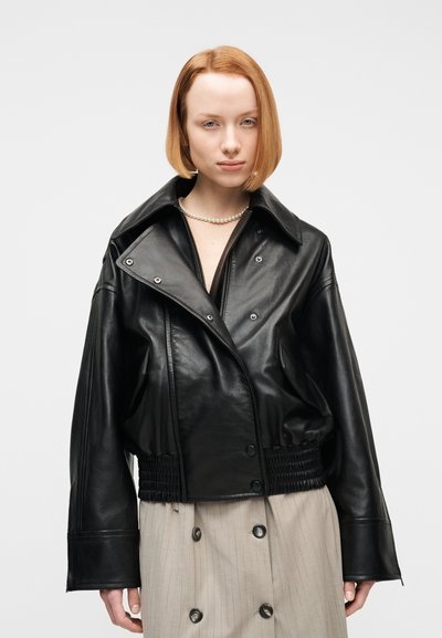 Νεαρή γυναίκα με ίσια κόκκινα μαλλιά, που φοράει μαύρο δερμάτινο μπουφάν, κολιέ με πέρλες και μπεζ διπλό κουμπωτό φούστα, σε φωτεινό φόντο.