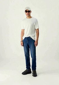 Bela kratka majica s kratkimi rokavi, modri jeans, črne teniske in siva kapa. Nosim sončna očala in stojim pred nevtralno ozadjem.